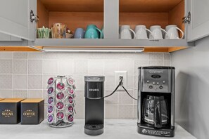 Una cocina con cafetera, azúcar y cajas de café, y un escurreplatos para tazas.