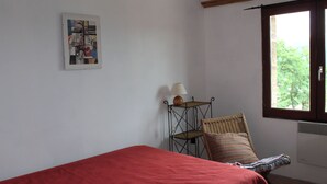 Een kamer met een bed, een stoel, een tafel met een lamp, een foto aan de muur en een raam met uitzicht.