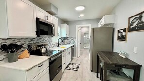 A compact kitchen with white cabinets, a microwave, and a sink. There is a small dining table with a placemat. - Mangrove Nest (Fort Myers Beach)