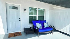 A white door with a glass panel, a blue sofa with patterned cushions, a small black bench, and a 'hello' mat on the porch. - Mangrove Nest (Fort Myers Beach)