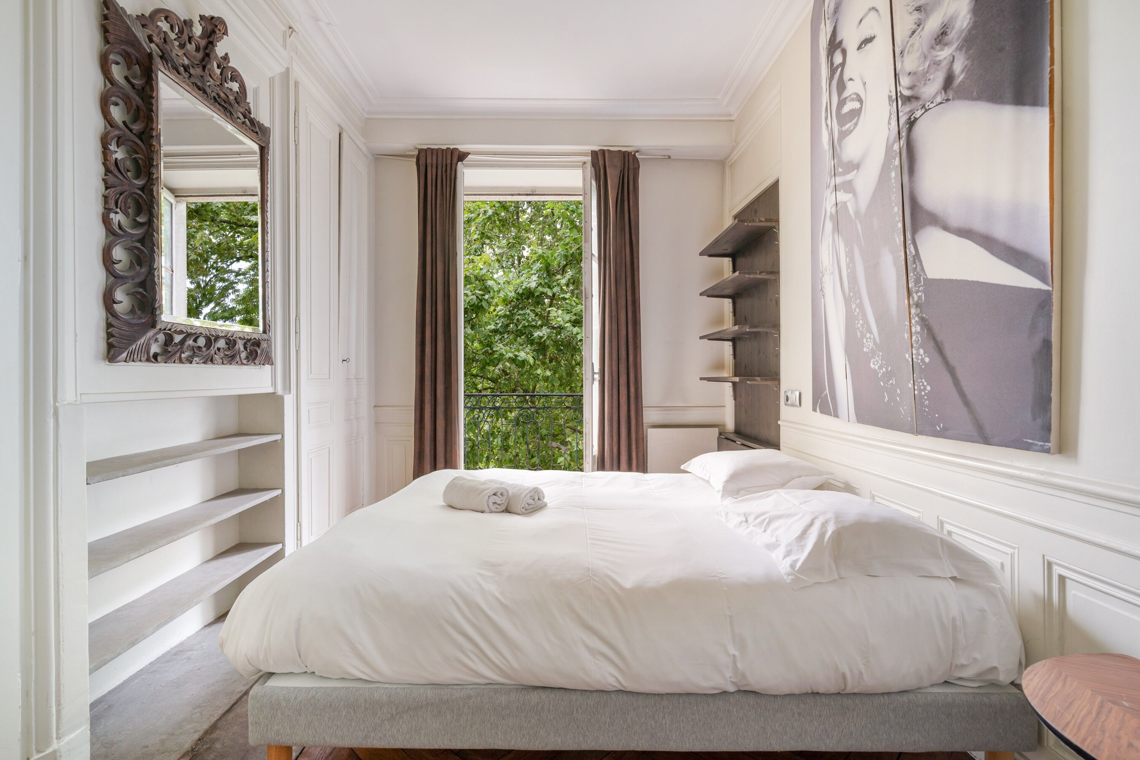 Une chambre d’hôtel avec un grand lit, un miroir, une fenêtre avec des rideaux et un mur orné d’un grand tableau encadré.