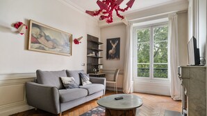 Ein Wohnzimmer mit grauem Sofa, einem runden Couchtisch, einem Bild eines Rehs und einem Fenster mit Blick auf Bäume.