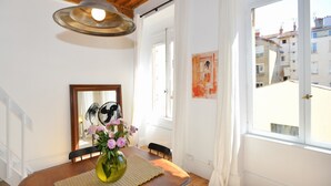 Uma sala de jantar com uma mesa de madeira, um vaso com flores, um espelho e uma janela com vista para prédios.