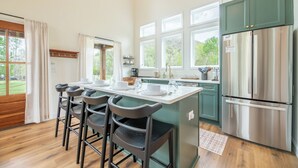 A modern kitchen with a central island, bar stools, a stainless steel refrigerator, and large windows. - Gorgeous 1BD+Loft Cabin w/ River Access + Fireplace (Townsend)