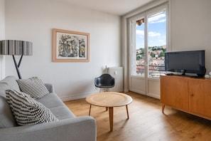 Ett vardagsrum med en grå soffa, ett runt träbord, en svart stol, ett inramat konstverk, en tv på ett träskåp och en utsikt över byggnader genom ett stort fönster.