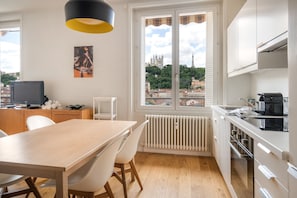 Une cuisine moderne avec des meubles blancs, un coin repas doté d’une table et de chaises en bois, une vue sur le paysage urbain par une fenêtre et un luminaire suspendu noir et jaune.
