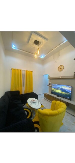 Una sala de estar con sofá, un sillón amarillo, una mesa de centro, televisor y lámpara de techo.