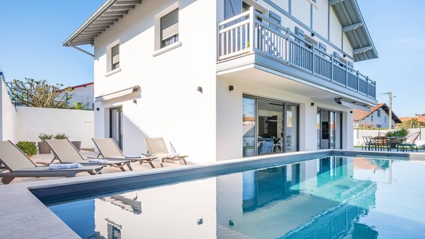 Una casa moderna de dos pisos con piscina, sillas de descanso y un balcón.