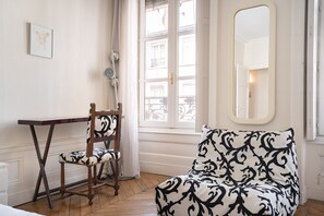 Una habitación con una silla de madera, un sofá estampado, un espejo y una ventana con vistas a edificios.