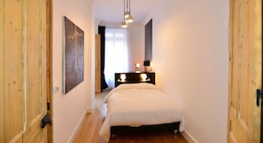 Un pasillo estrecho que conduce a una cama con cabecera, un cuadro en la pared y una mesita de noche con lámparas.