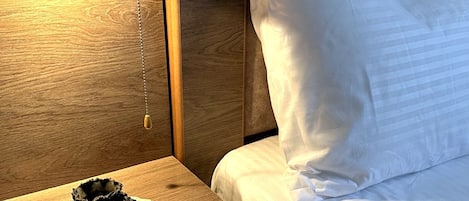 Una cama bien hecha con sábanas blancas, una mesita de noche de madera con un adorno pequeño y una lámpara de pared que proporciona luz.
