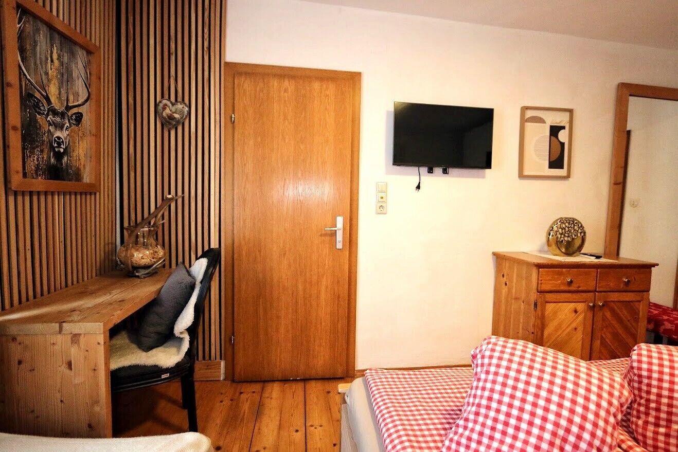 Ein Zimmer mit Holztisch, Stuhl, Bett mit karierter Bettwäsche, Holzschrank, Fernseher und einem wandmontierten Spiegel.