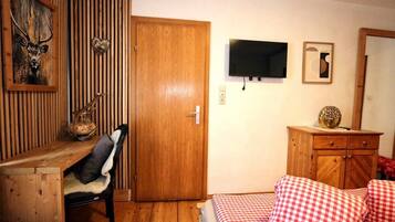 Ein Zimmer mit Holztisch, Stuhl, Bett mit karierter Bettwäsche, Holzschrank, Fernseher und einem wandmontierten Spiegel.