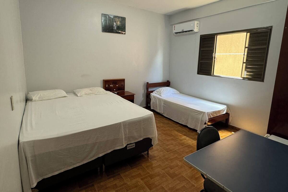 Una habitación con dos camas, una mesita, aire acondicionado y una ventana con persianas.