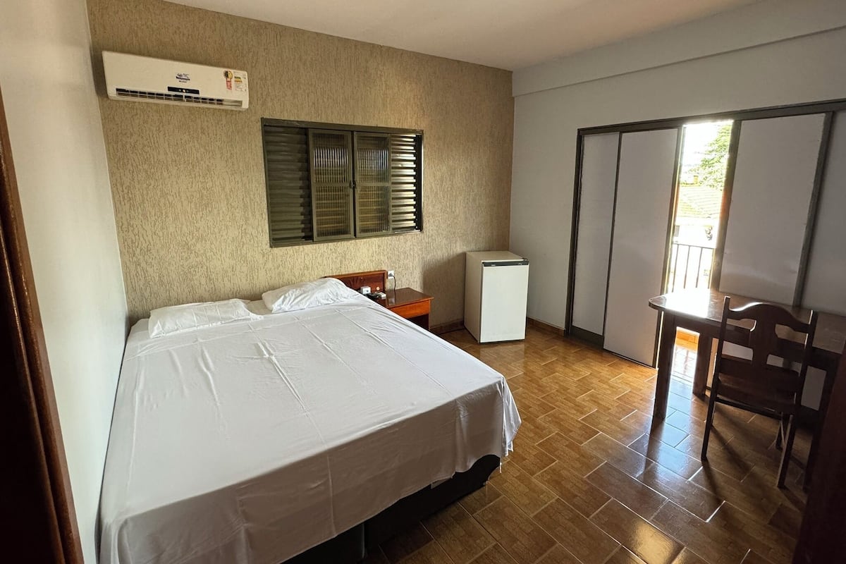 Habitación de hotel con cama, una mesita, una silla, refrigerador y puerta corrediza que da a un espacio al aire libre.