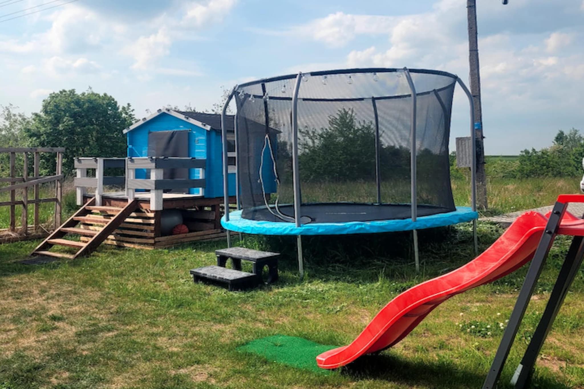 Ein Trampolin mit Sicherheitsnetz, eine Rutsche, ein kleines blaues Spielhaus und eine Holztreppe, die zum Spielhaus führt.
