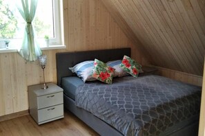 Une chambre avec un plafond en pente, un lit avec une literie à motifs, une table de chevet avec des tiroirs et une fenêtre avec un rideau.