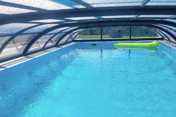An indoor swimming pool with clear blue water, a green inflatable float, and a view of the outside through a glass ceiling.