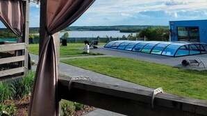 On peut voir une serre en verre, une terrasse en bois et un bâtiment bleu, avec un lac en arrière-plan.