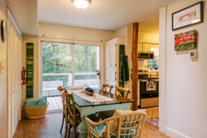 A dining area with a table and chairs, a kitchen in the background, and a view of trees through a sliding door. - the Bird House/Center of Ocean Springs (Ocean Springs)