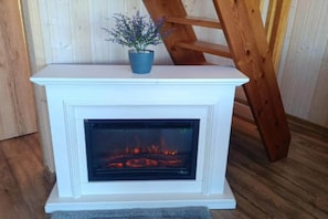 Una chimenea blanca con una maceta azul encima, una escalera de madera apoyada y fuego ardiendo en su interior.
