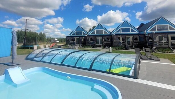 Een zwembad met een glazen overkapping, een houten terras en een rij gebouwen met blauwe daken in de achtergrond.