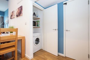 Ein kleines Zimmer mit Waschmaschine, Tisch mit Stühlen und Kleiderschrank mit blauem Akzent.