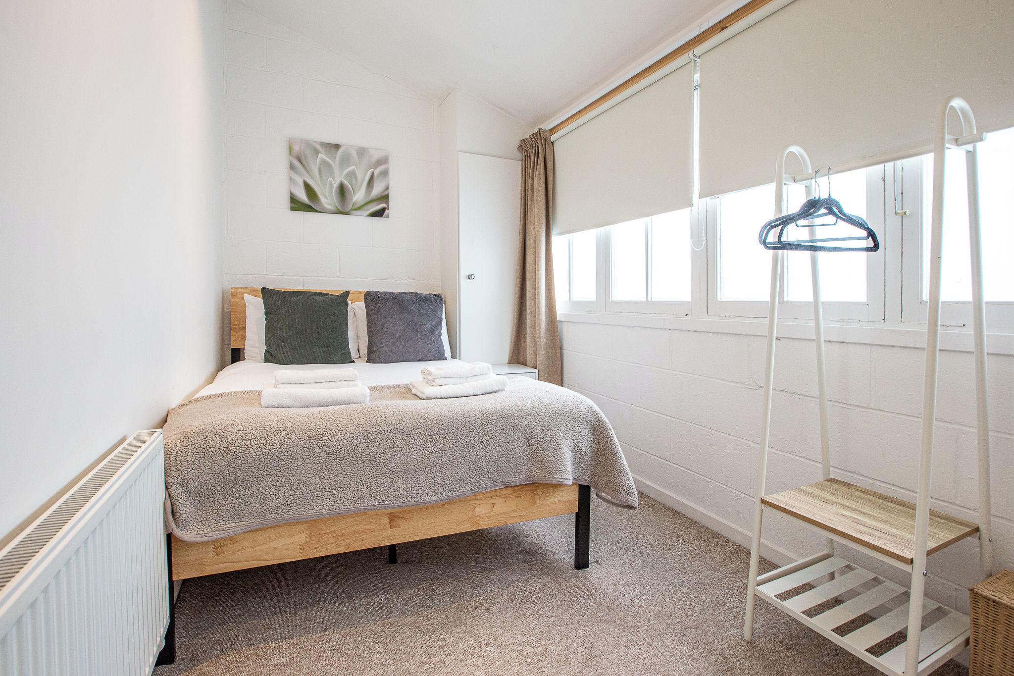 A bedroom with a bed, a radiator, a window with curtains, a wall-mounted shelf, and a hanging clothes rack.