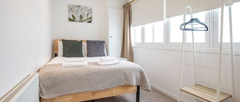 A bedroom with a bed, a radiator, a window with curtains, a wall-mounted shelf, and a hanging clothes rack.