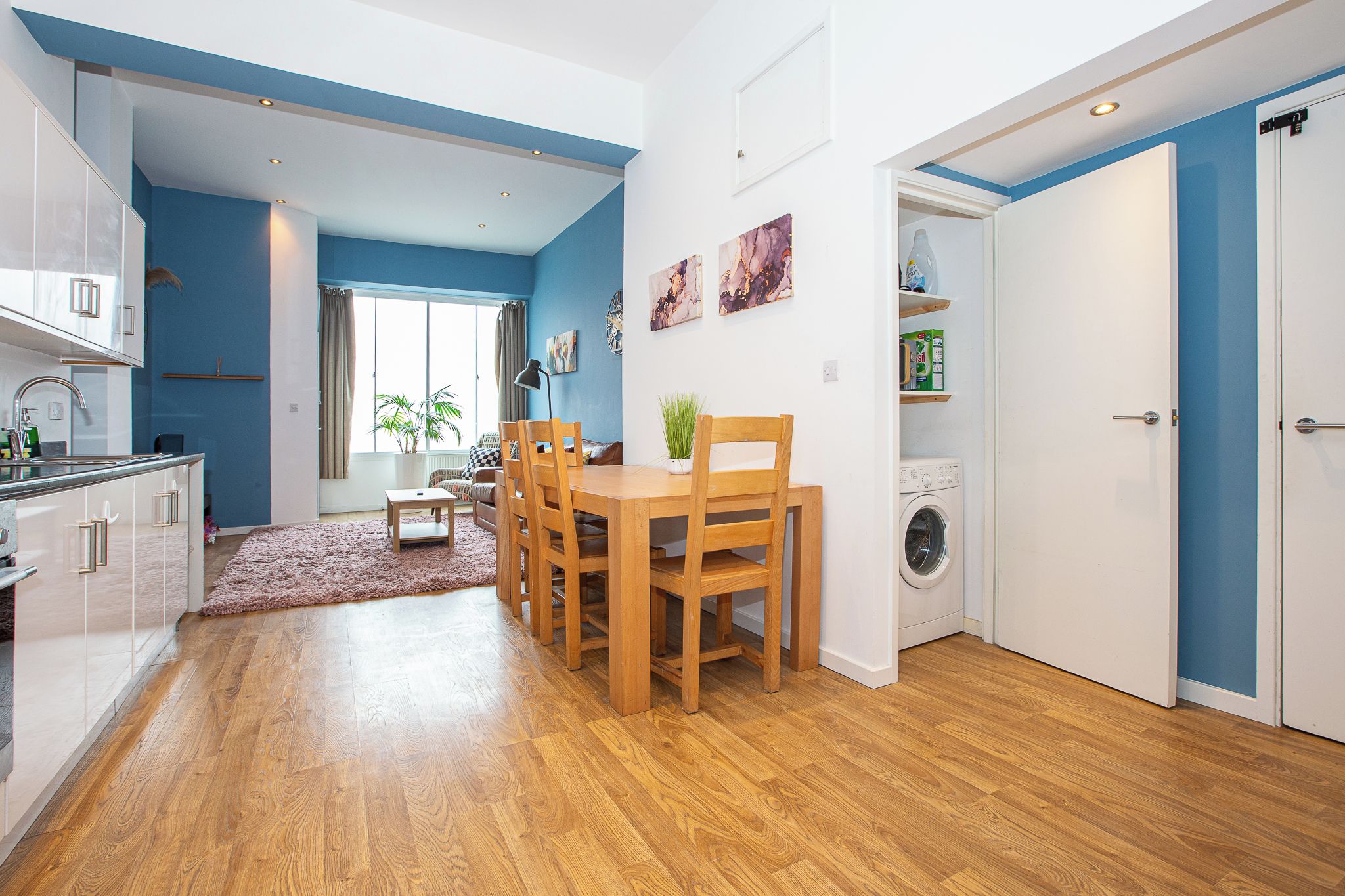 A modern kitchen with white cabinets, a dining area with wooden chairs, a washing machine, and a blue accent wall.