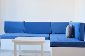 A blue sofa with cushions and a small table in a room with white walls and tiled flooring.