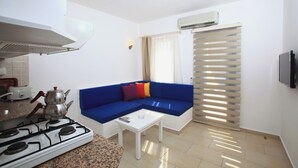 Un espacio habitable compacto con kitchenette, un sofá azul, una mesita y un televisor montado en la pared.