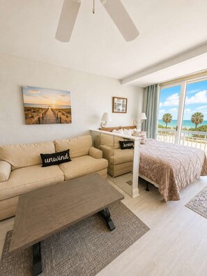 A hotel room with a bed, a sofa, a desk, a view of the beach, and a painting on the wall. - Oceanfront Pool View Condo by Pier! Yachtsman S205 (Myrtle Beach)