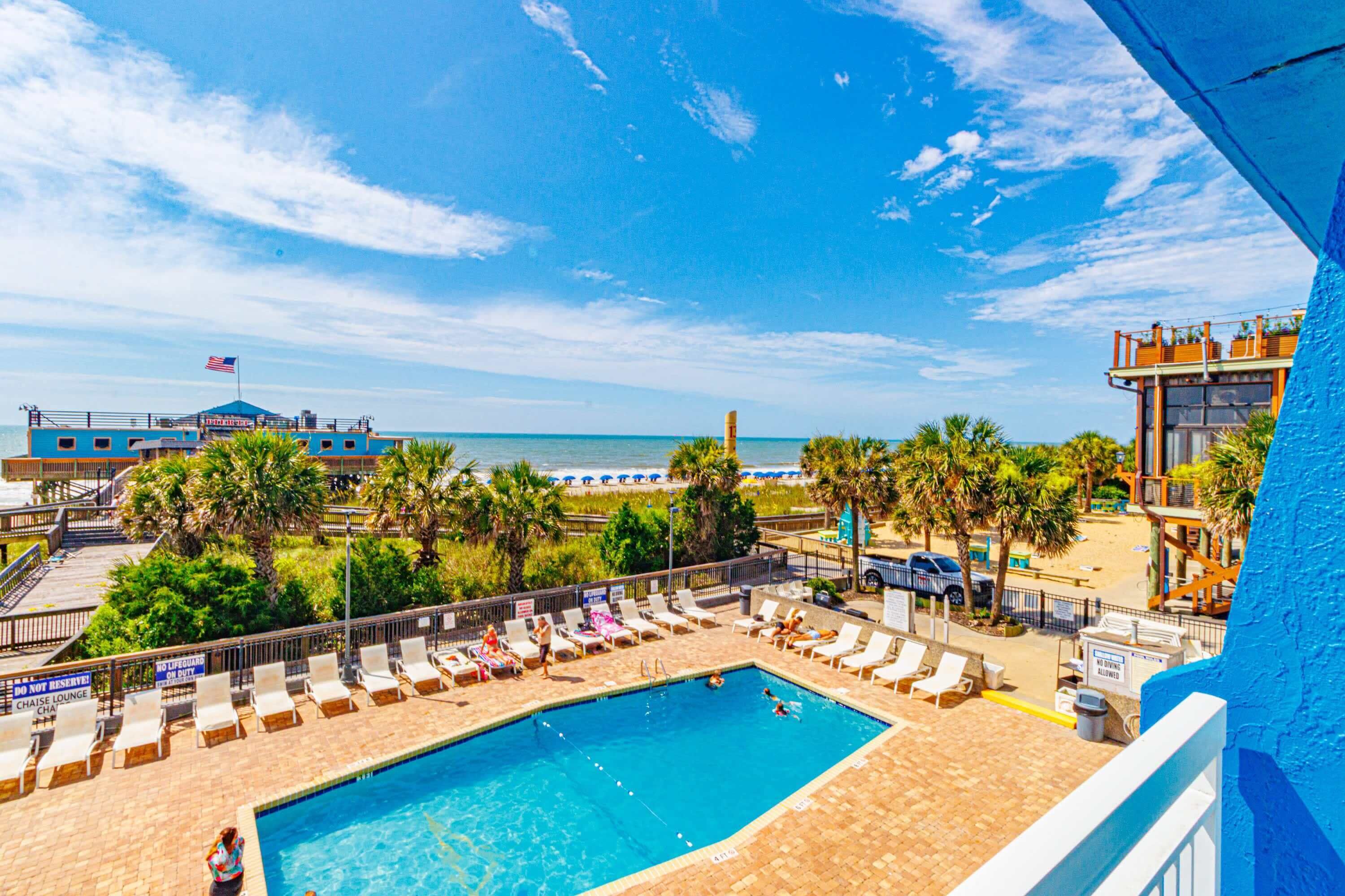 A pool area with lounge chairs, a lifeguard tower, and a view of the beach.