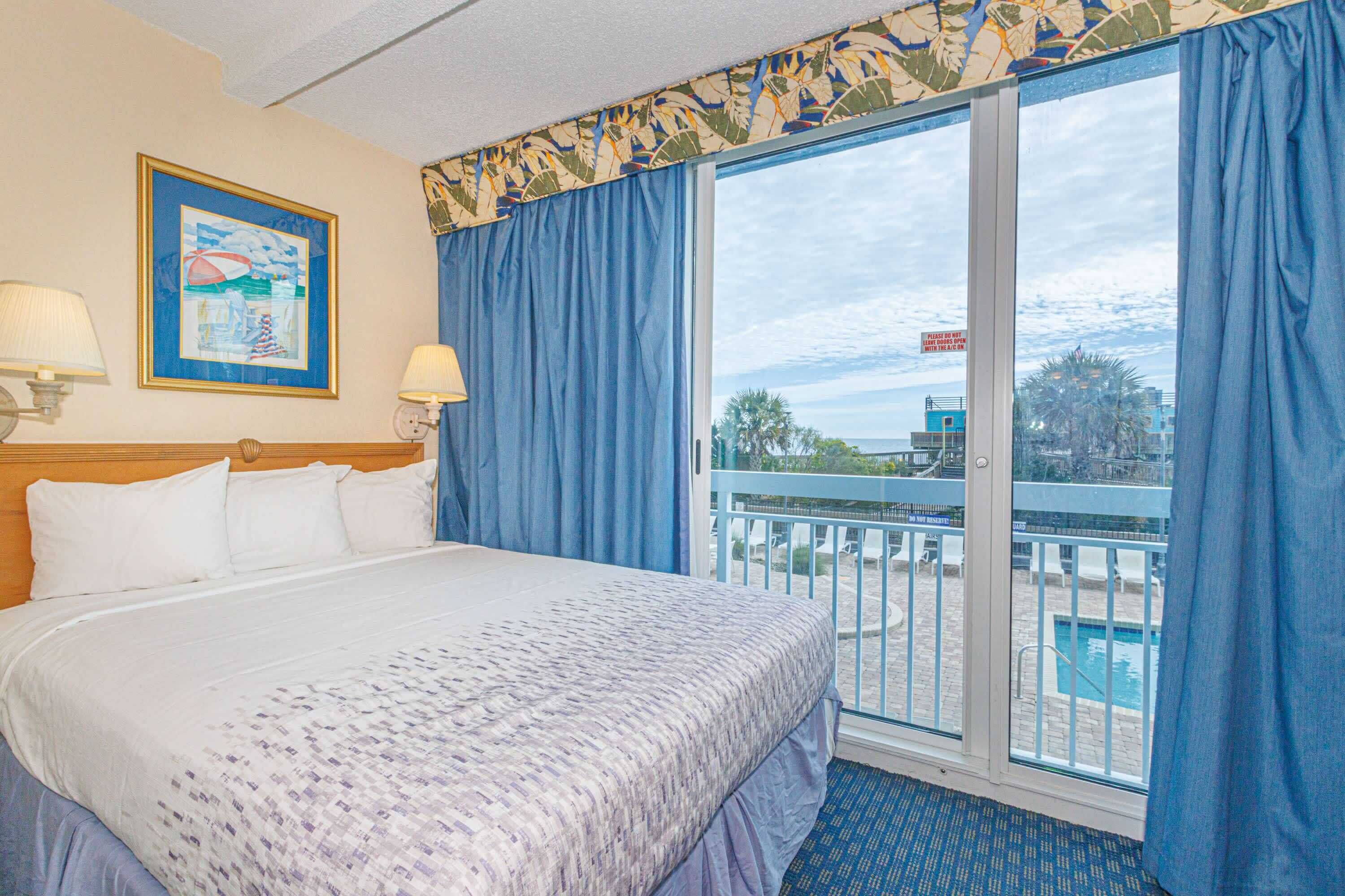 Una camera d'hotel con un letto, due lampade, un quadro e un balcone con vista sulla piscina.