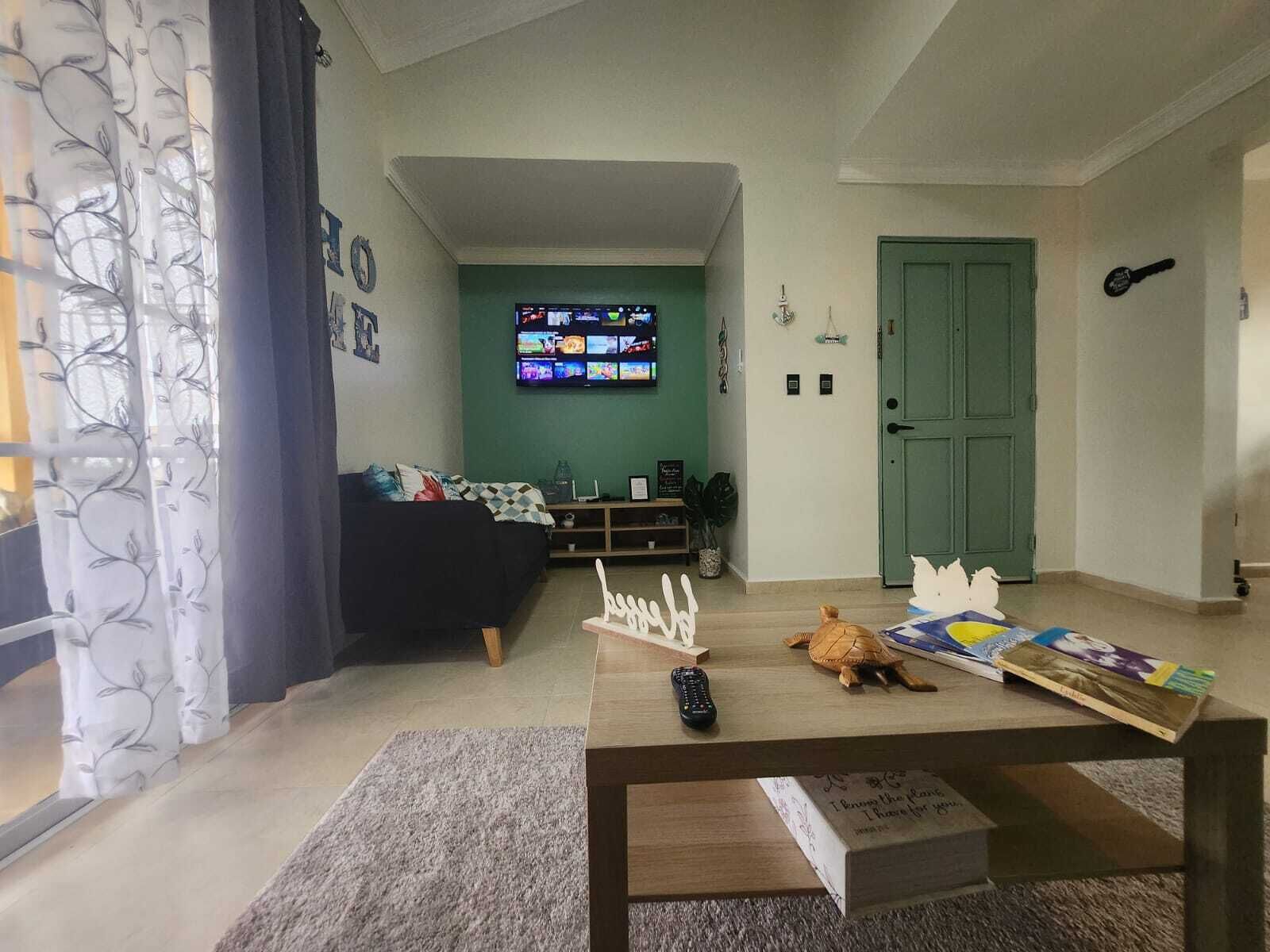 Una sala de estar con un sofá, una mesa de centro con un libro y un control remoto, un televisor en la pared y una puerta verde.