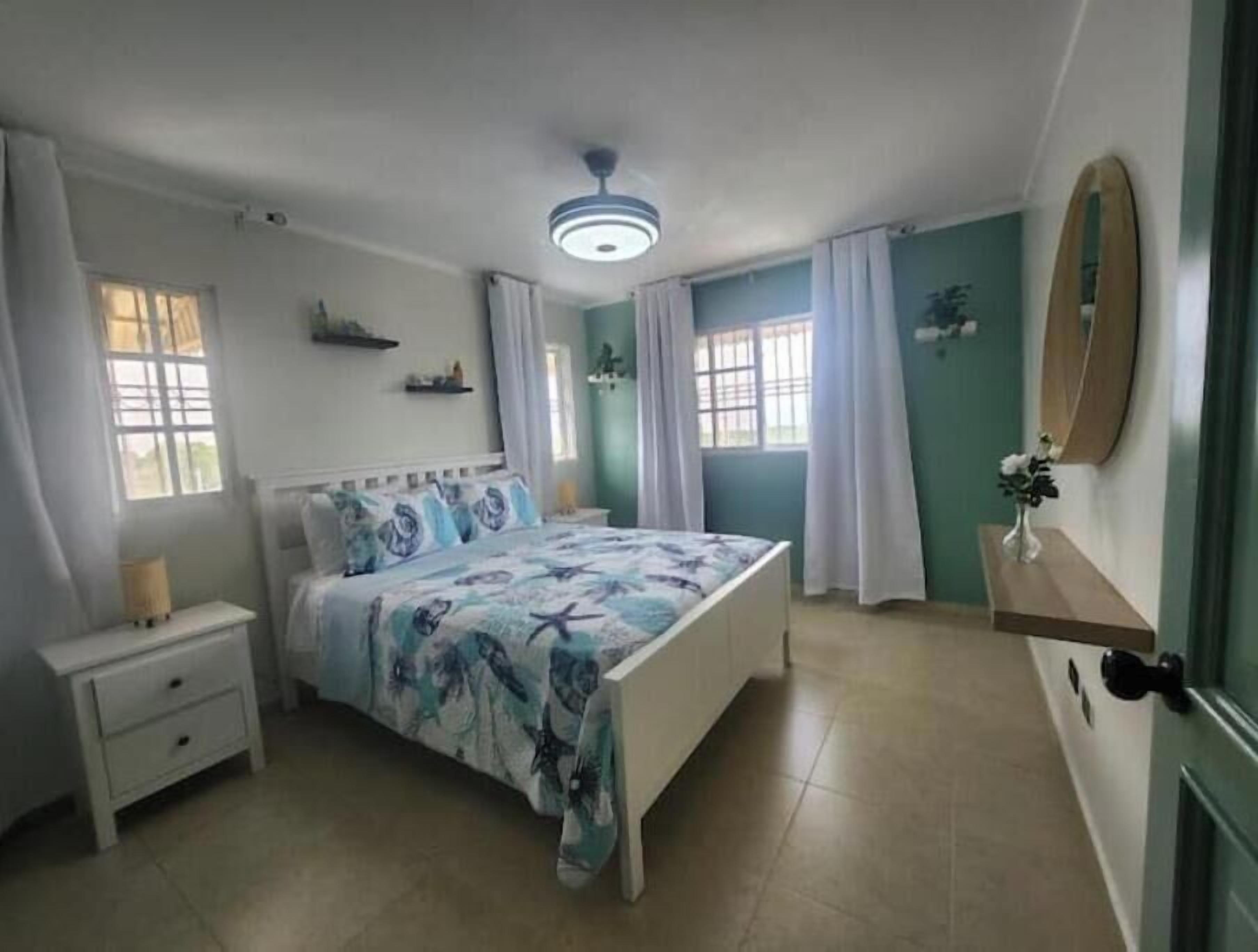 A bedroom with a bed, bedside tables, a ceiling fan, windows with curtains, a mirror, and a shelf with a vase.