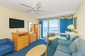 Quarto de hotel com televisão, ventoinha no teto, sofá, escrivaninha, cadeira, cómoda e vista para o oceano.