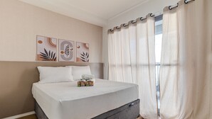 A neatly made bed with a grey headboard, a window with sheer curtains, and two wall art pieces above the bed. - Your Code Liberdade (São Paulo)