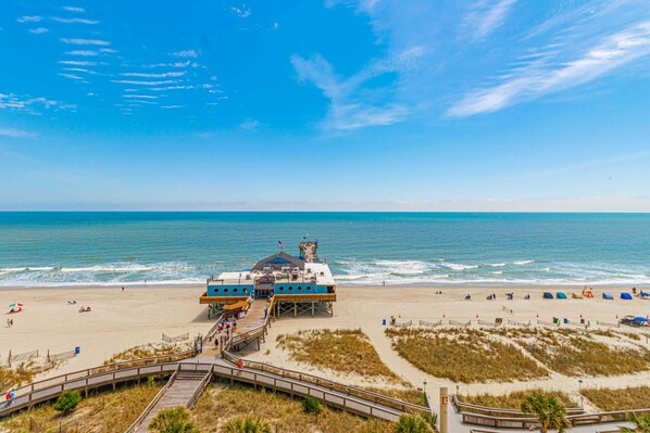 A beachside building with a pier, a lifeguard tower, and a sandy beach with umbrellas.