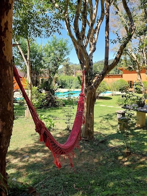 A hammock hanging between trees in a garden with a pool and a building in the background.