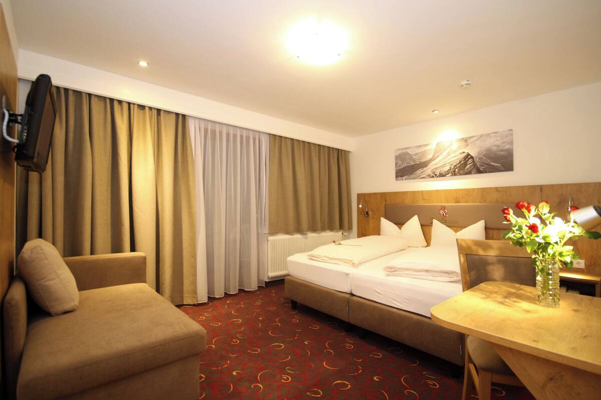 A hotel room with a bed, a sofa, a table, a vase with flowers, a painting on the wall, and a television mounted on the wall.