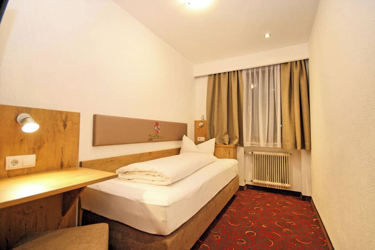A hotel room with a bed, a desk, a chair, a window with curtains, and a radiator.