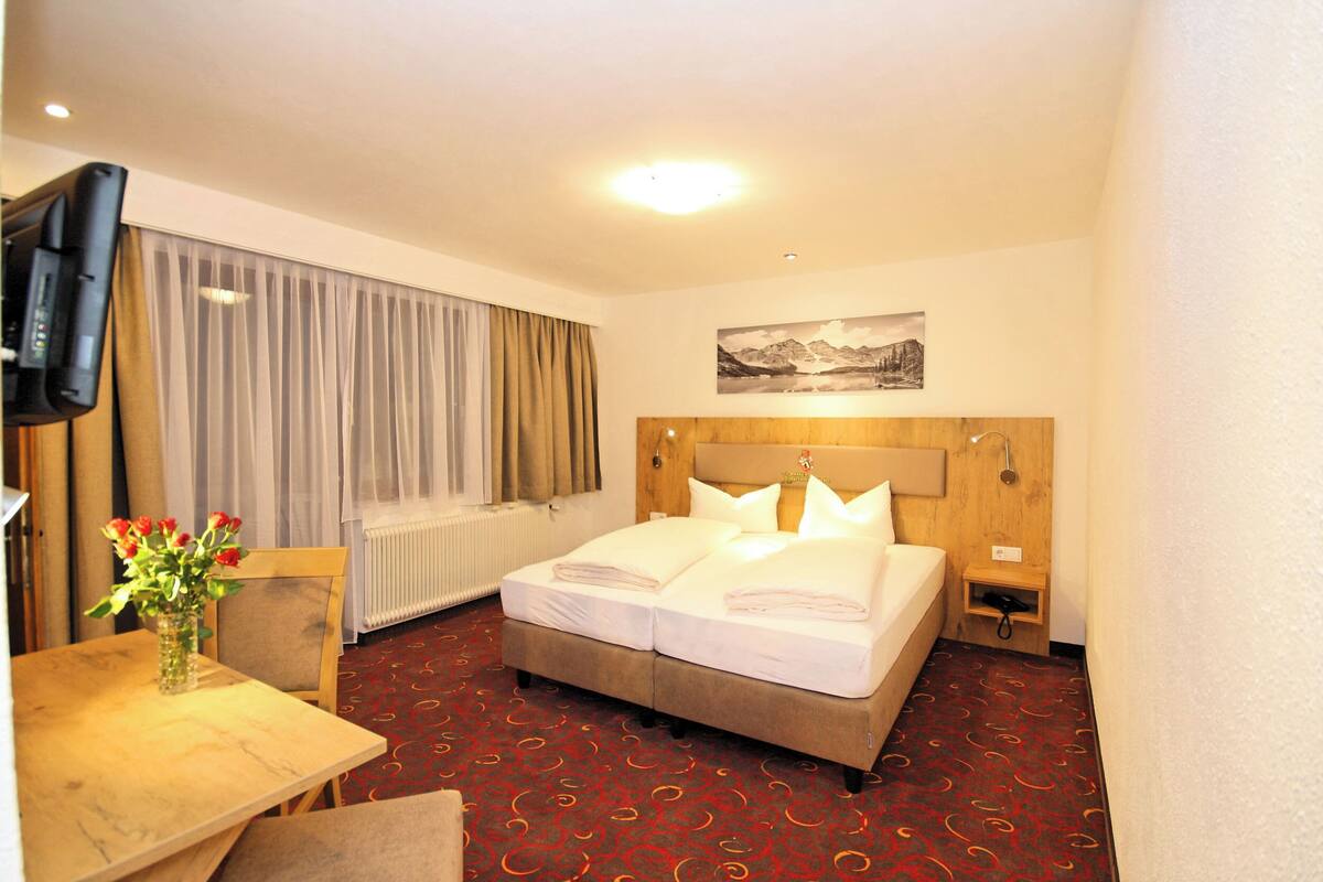 A hotel room with a bed, a TV, a table with a vase of flowers, and a painting on the wall.