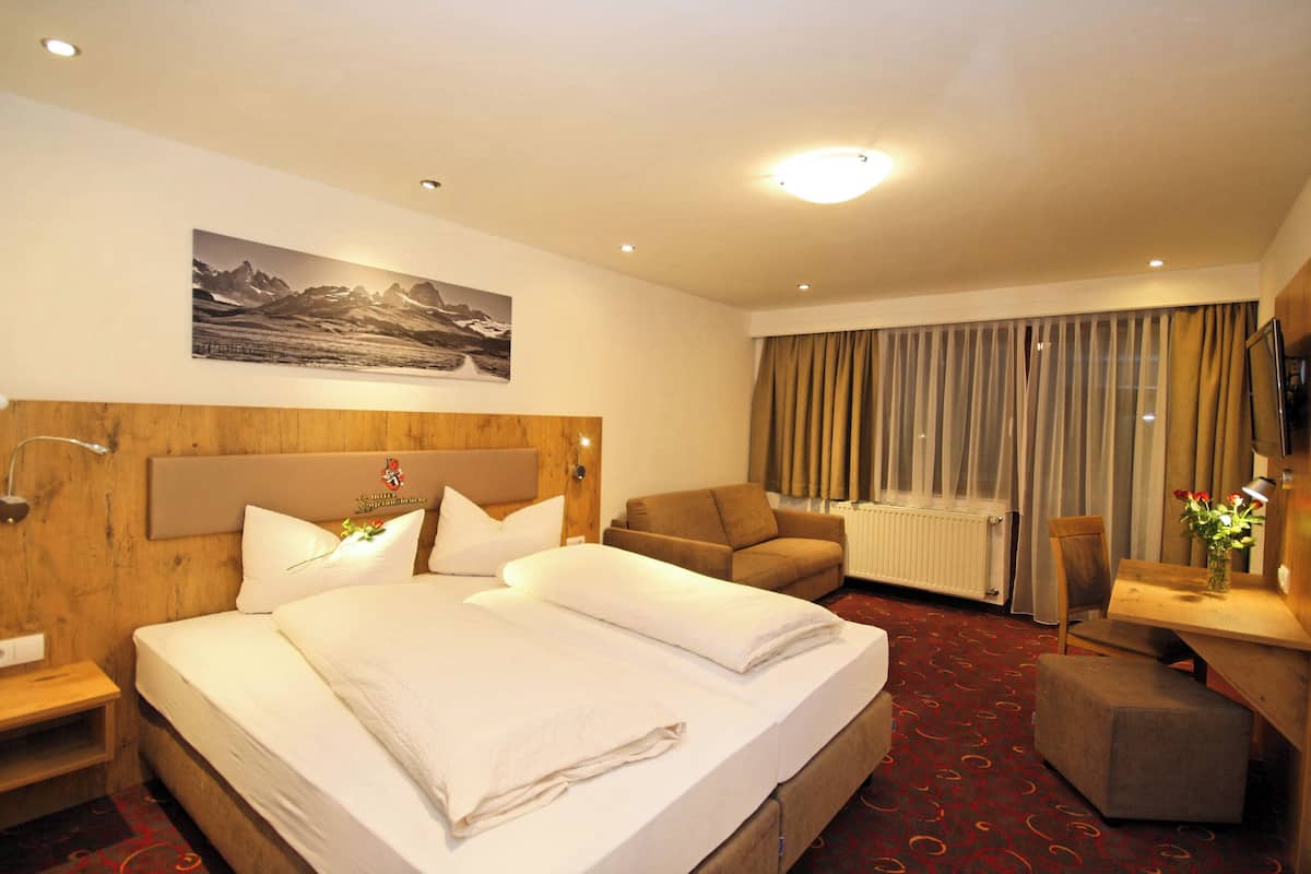 A hotel room with a large bed, a wooden headboard, a desk with a chair, a window with curtains, and a painting on the wall.