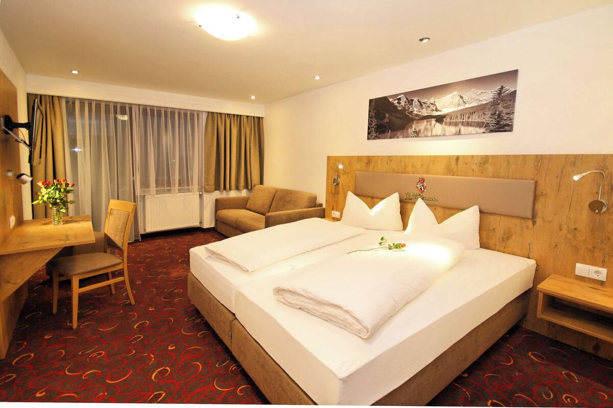 A hotel room with a large bed, a desk, a chair, a wooden headboard, and a wall-mounted picture.
