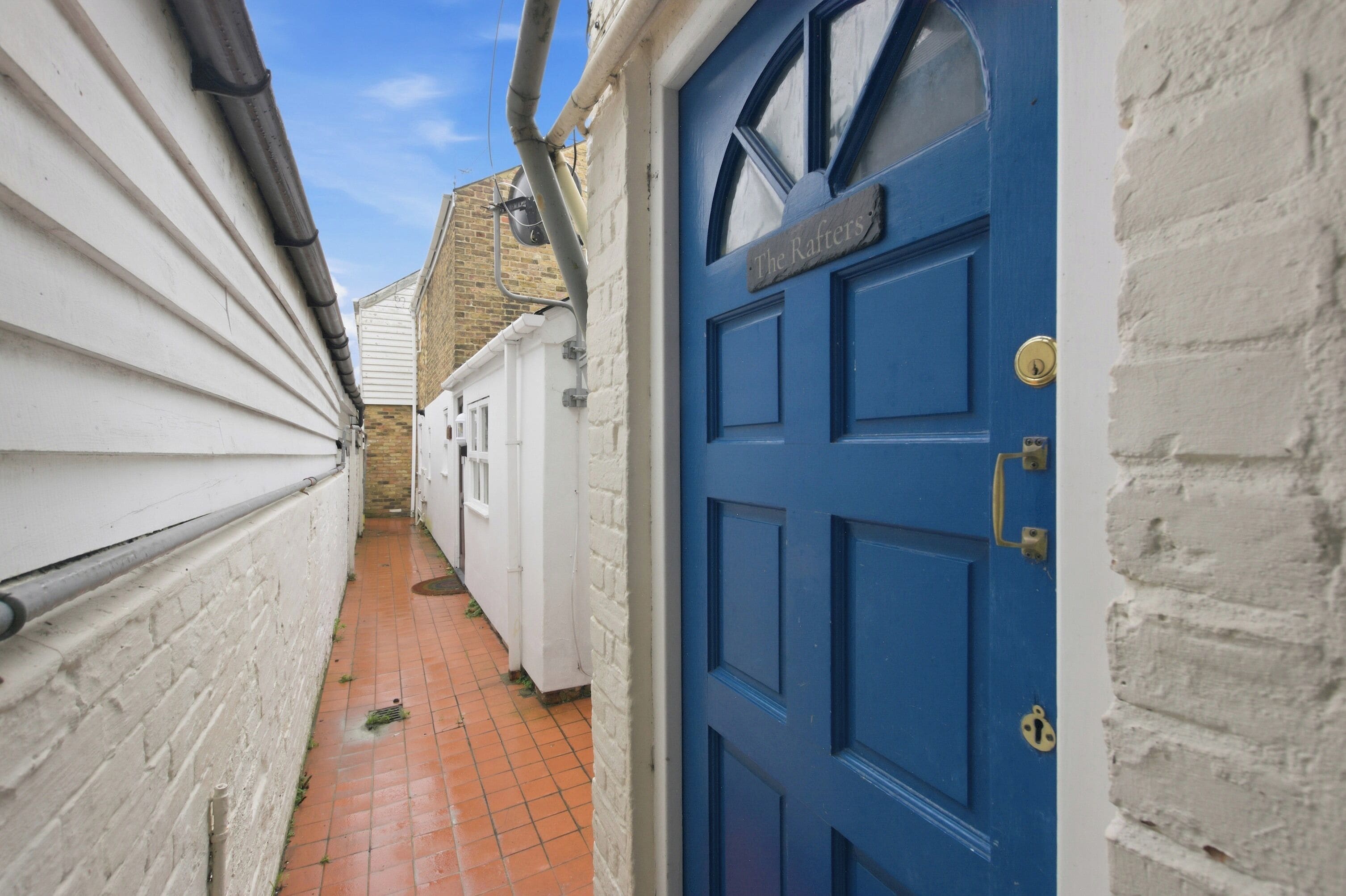 A narrow alleyway with a blue door labeled 'The Rafters' and a white brick wall.