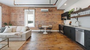 Une cuisine moderne avec un mur en briques, un coin repas avec une table et des chaises blanches, et un espace de vie comprenant un canapé et une table basse.