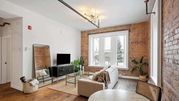 Un salon moderne avec un mur en briques, une grande fenêtre, un téléviseur à écran plat, un canapé, une table basse et une table à manger.