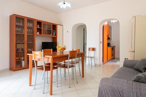 Un salon moderne avec un coin repas, une cuisine ouverte sur le salon par une arche, et un canapé gris.